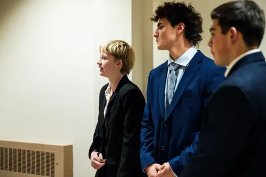 Three people in business attire stand indoors, facing left. One person has short blonde hair, and the other two have dark hair. They appear attentive and are possibly in a professional or formal setting.