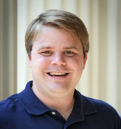 A smiling person with short, light brown hair wearing a navy blue polo shirt stands in front of a neutral background.