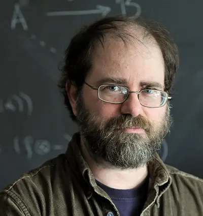 A bearded man with glasses stands in front of a chalkboard covered with diagrams and mathematical symbols. He has a contemplative expression and wears a brown shirt over a black t-shirt.