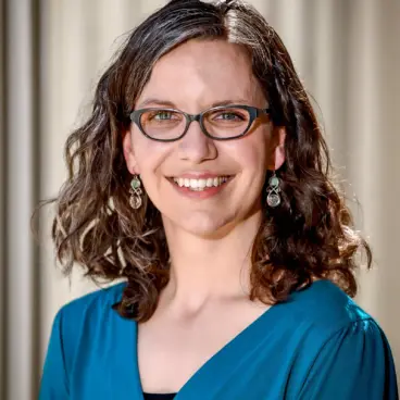 A woman with shoulder-length curly hair and glasses smiles at the camera. She is wearing a teal top and dangling earrings, standing against a neutral background.