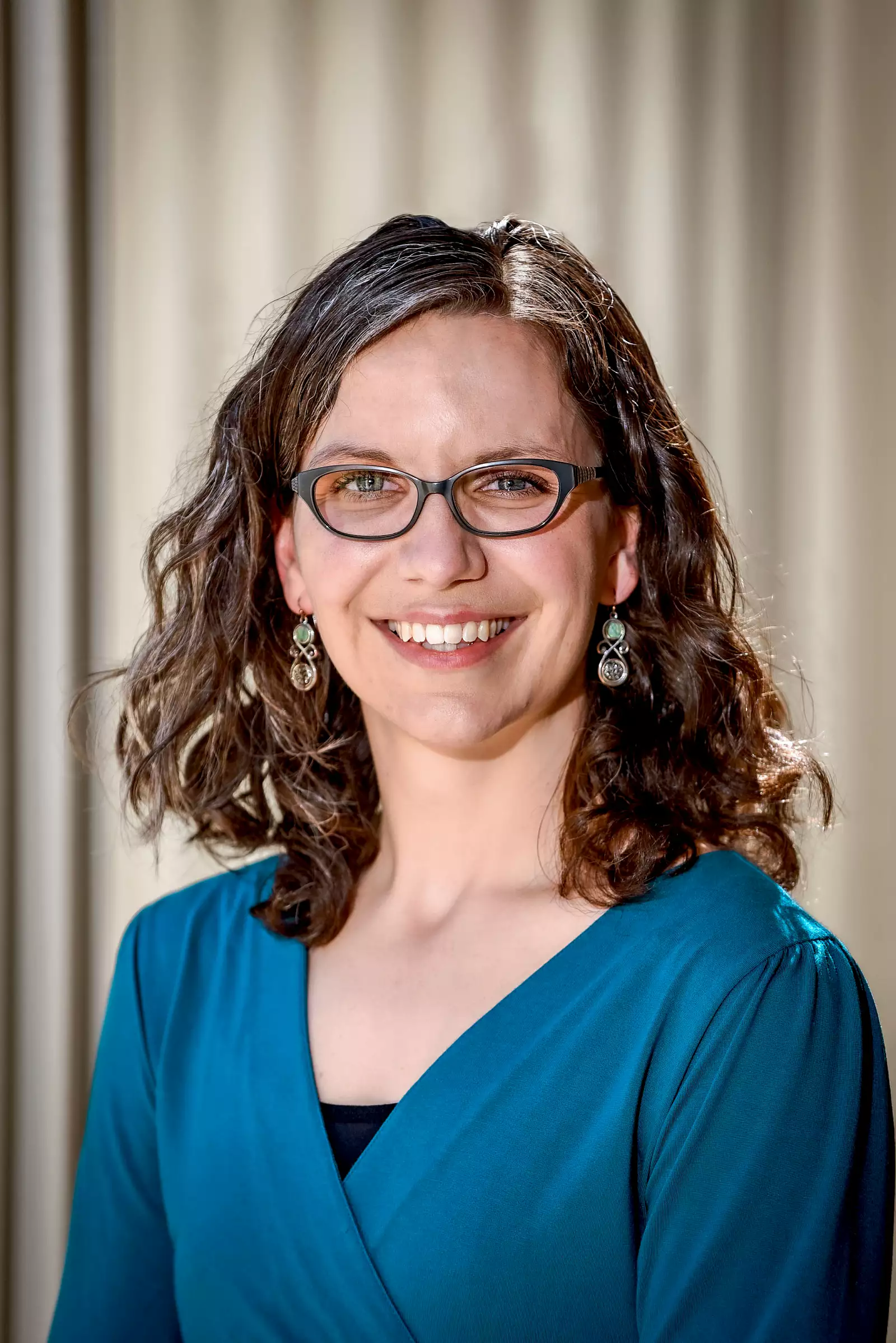 A woman with shoulder-length curly hair and glasses smiles at the camera. She is wearing a teal top and dangling earrings, standing against a neutral background.