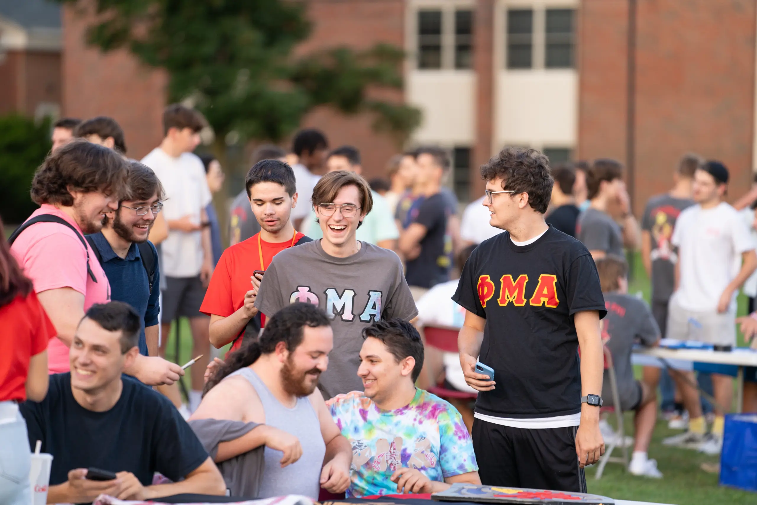 A group of young men gathered outside at a fraternity event. Some are standing, wearing fraternity-themed shirts, and engaging in conversation. Others are sitting, smiling, and interacting with each other. The background shows a building and greenery.