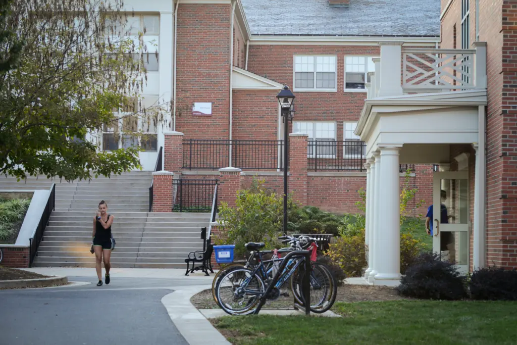 A person walks on a pathway in front of a brick academic building. Two bicycles are parked nearby, and there are steps leading up to the entrance. Trees and shrubs are visible, enhancing the campus atmosphere.