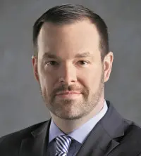 A man in a suit and tie with short, dark hair and a beard is looking at the camera. The background is a plain, neutral color.