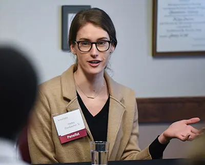 A person wearing glasses and a beige coat speaks during a panel discussion. A name tag reading "Panelist" is visible on their coat. The background has a framed document on the wall.