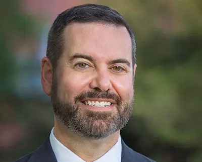 A man with a beard smiling, wearing a suit and tie. The background is blurred with green and brown tones.