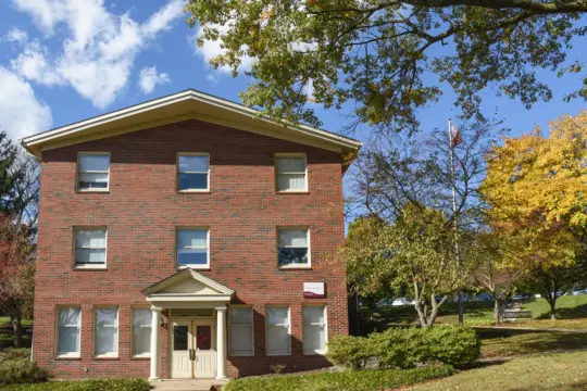 A three-story brick building with a peaked roof, surrounded by trees with autumn foliage. The sky is blue with a few clouds. A path leads to the entrance, which has a small overhang supported by columns.