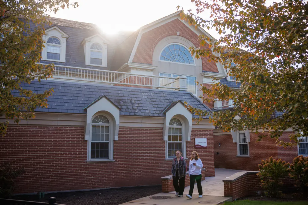 Two people walk on a path in front of a large brick building with a steep slate roof and arched windows. The sun shines brightly from behind the building, and trees with autumn leaves frame the scene.
