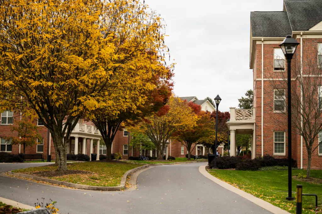 A scenic view of West Hall with red brick buildings and a winding path. Autumn trees with vibrant yellow and red leaves line the pathway under a cloudy sky. People are walking in the distance.