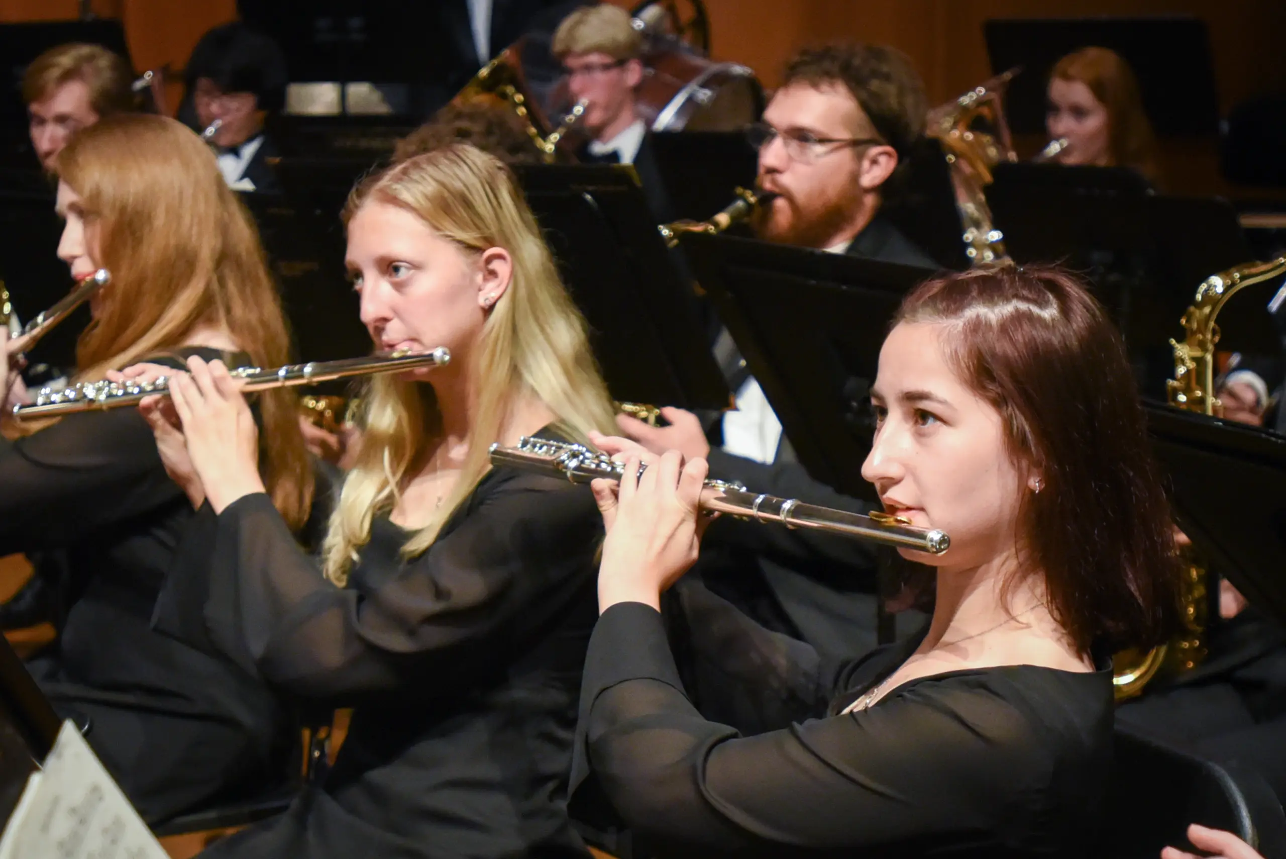 Musicians playing flutes in an orchestra. They are seated in rows, wearing black formal attire, and focusing on their sheet music. A variety of wind instruments are being played by other members in the background.