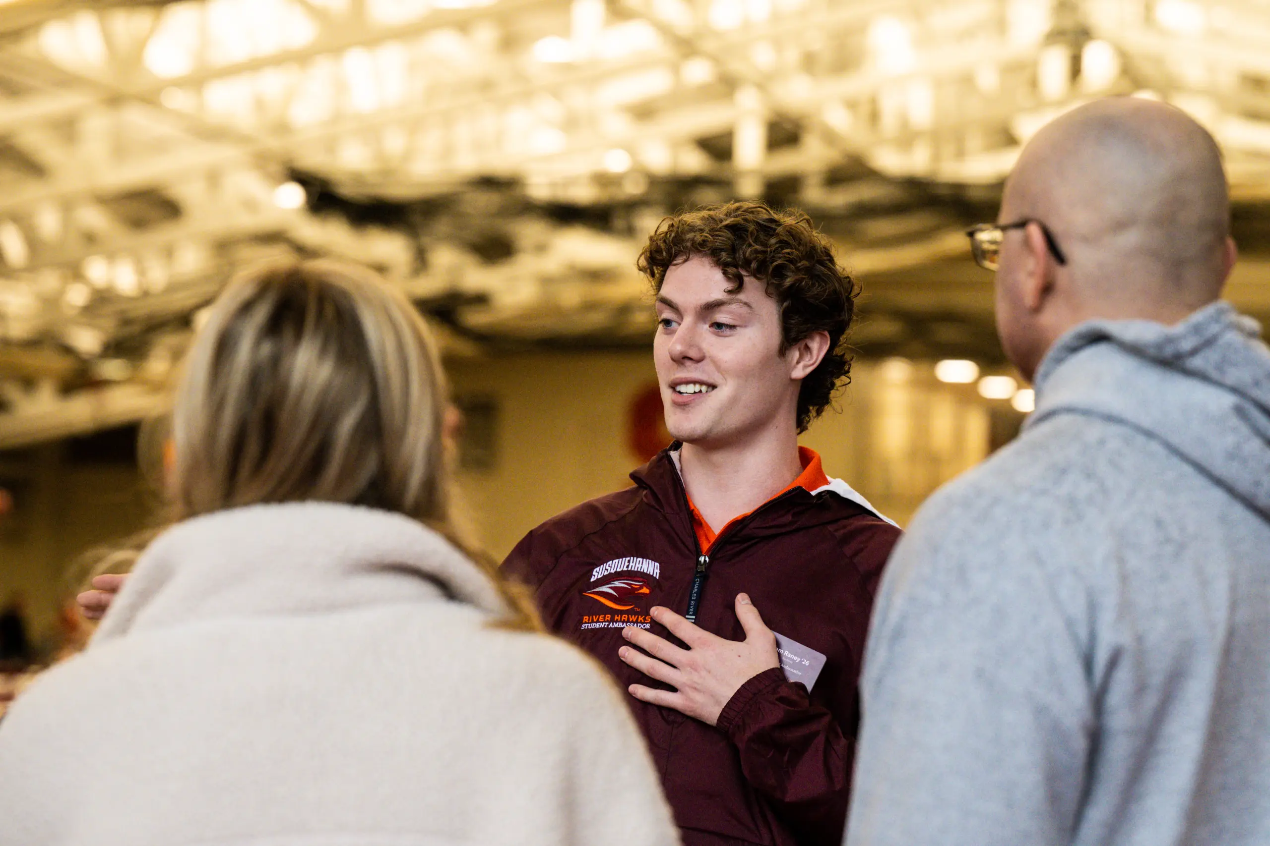 A young man wearing a maroon jacket gestures while speaking to two people in a casual setting. The background appears to be an indoor space with bright lighting and a high ceiling.