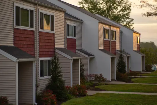A row of modern townhouses at sunset, featuring two-story structures with white and red paneled exteriors. Well-maintained lawns and small bushes line the pathway in front of the houses. Trees are visible in the background.