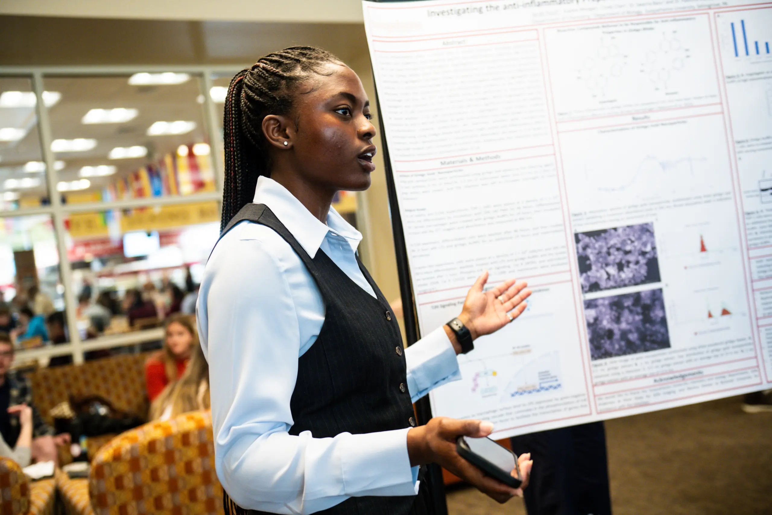 A person is giving a presentation in a formal setting, standing next to a research poster, embodying the "Learn By Doing" philosophy. They gesture with one hand while holding a phone in the other. Several people are seated in the background as large windows reveal a bright, busy room.