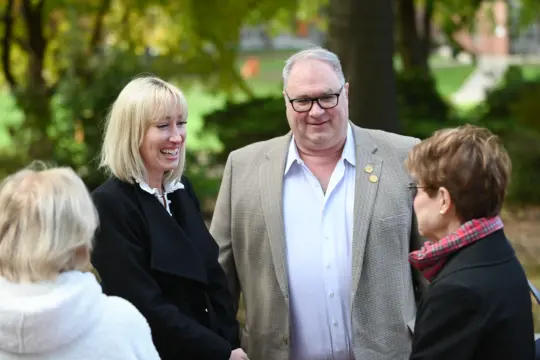 A group of four adults stands together outdoors, smiling and engaged in conversation. The background features greenery and trees that give the scene a serene ambiance. They appear relaxed and are dressed in warm clothing, suggesting a cool day.