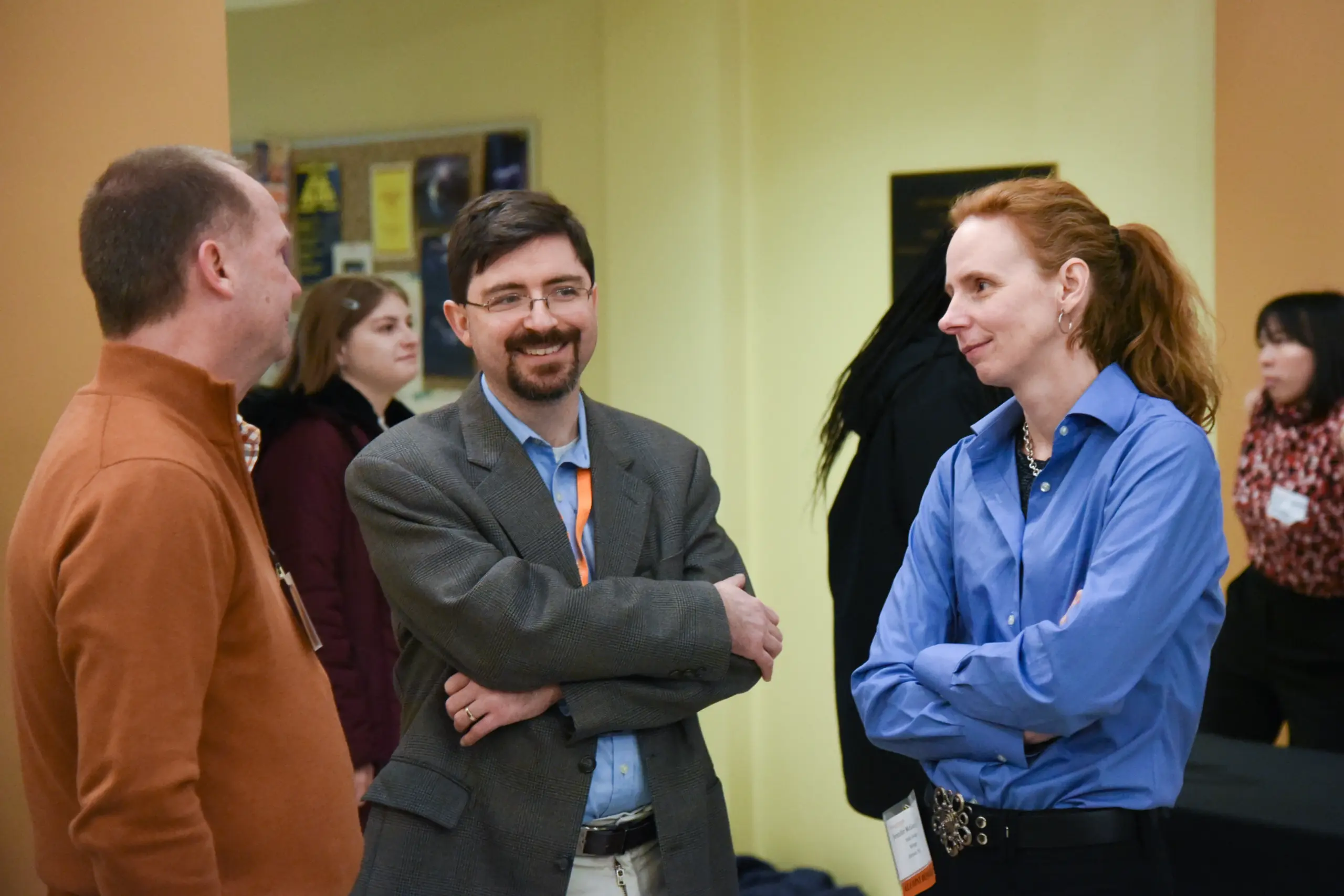 Three people are standing and talking in a room. One is wearing a gray blazer, another an orange sweater, and the third a blue shirt. They appear engaged in conversation. Other people are visible in the blurred background.