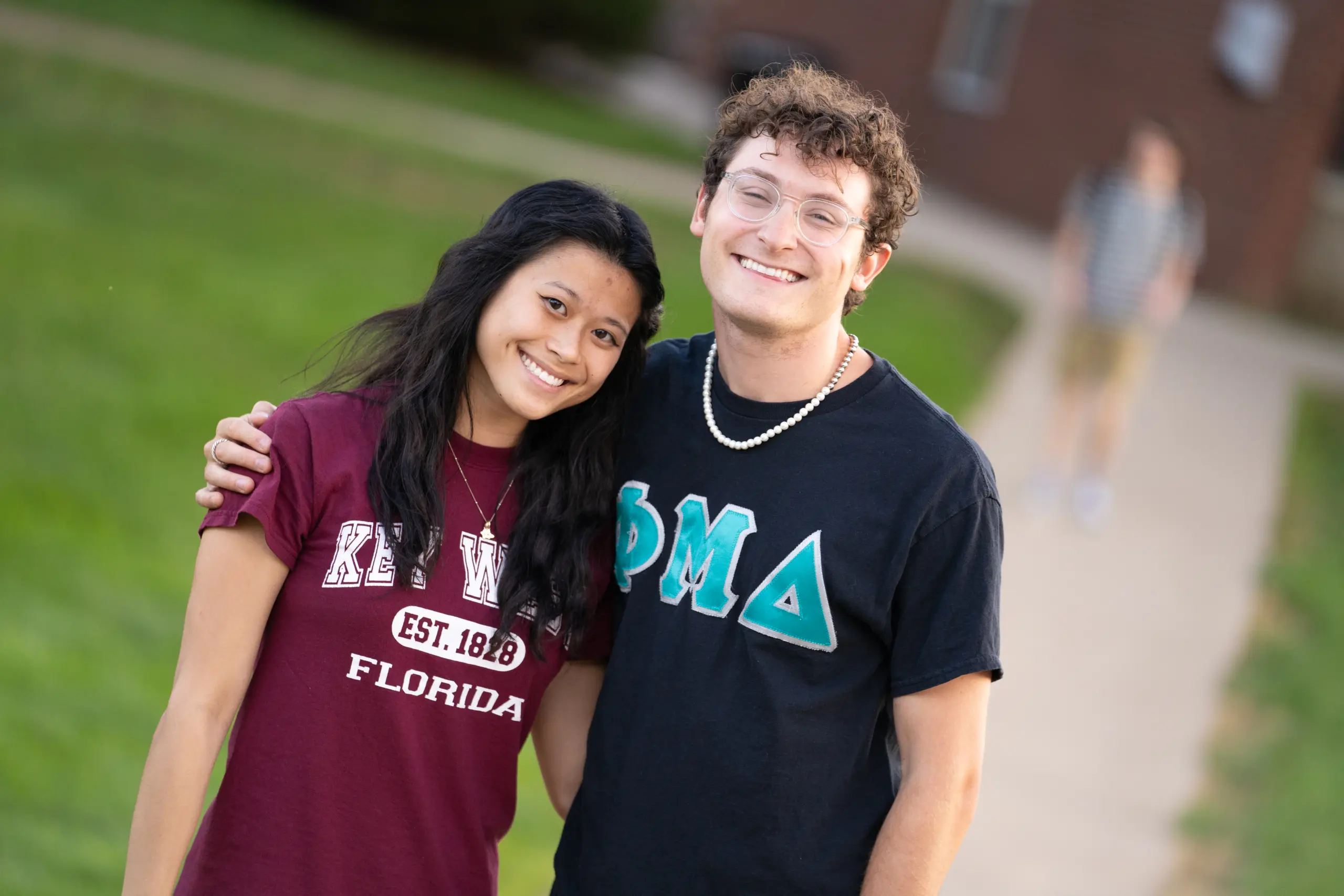 A young woman and man beam on a college campus; she's sporting a maroon "Key West Florida" shirt, and he's in a black Greek Life tee. The backdrop reveals a grassy area with a sidewalk where another student strolls by.