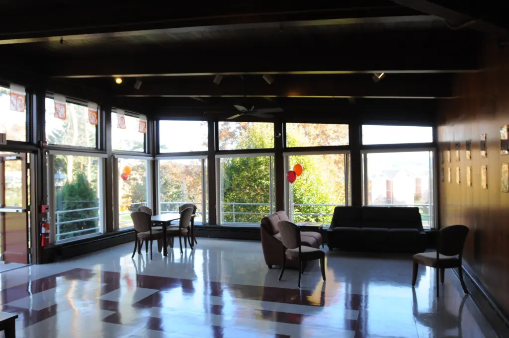 A sunlit room in an upper-level housing space features large windows, a few chairs, and tables arranged on a glossy floor. Balloons are tied to a chair, while a sofa rests against the windowed wall. Outside, trees and part of another building are visible.