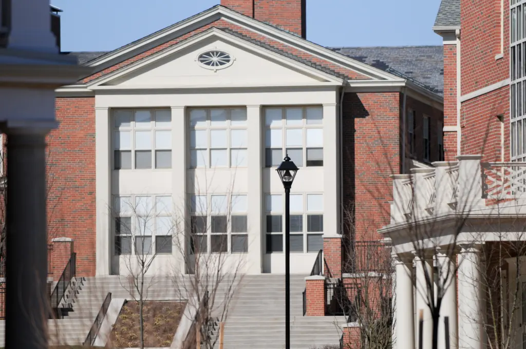 Front view of a classic red-brick building with a triangular pediment and large windows, characteristic of upper-level housing. Wide steps lead to the entrance, flanked by a black lamppost and a partially visible columned porch in the foreground.