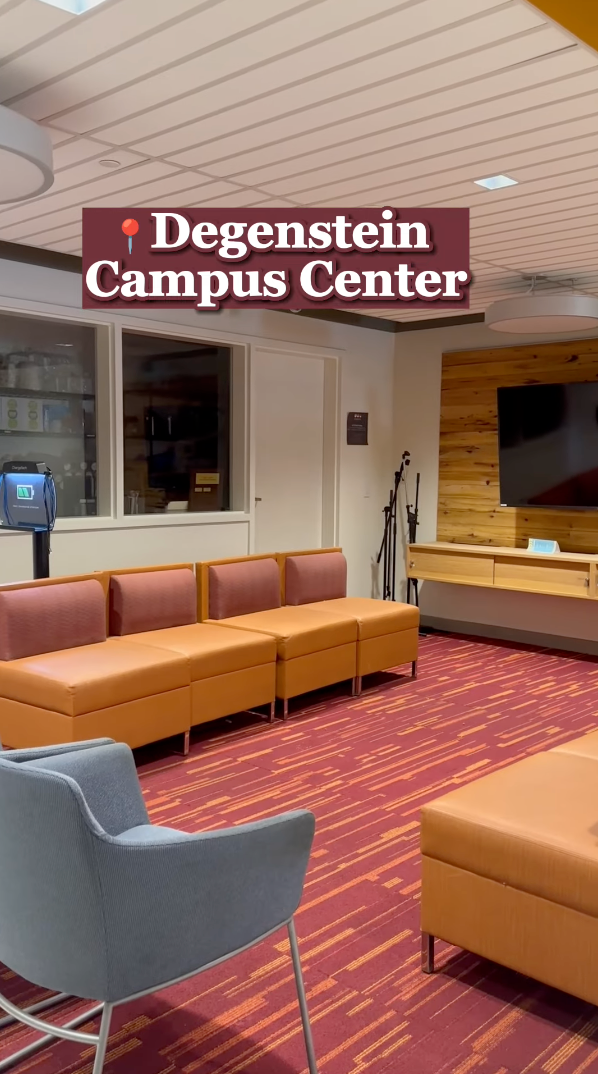 The modern lounge area inside Degenstein Campus Center seamlessly blends academics with relaxation, featuring orange couches, a gray armchair, a large flatscreen TV on a wooden wall, and a carpet with orange and red patterns. A water cooler and tripod stand are visible in the background.