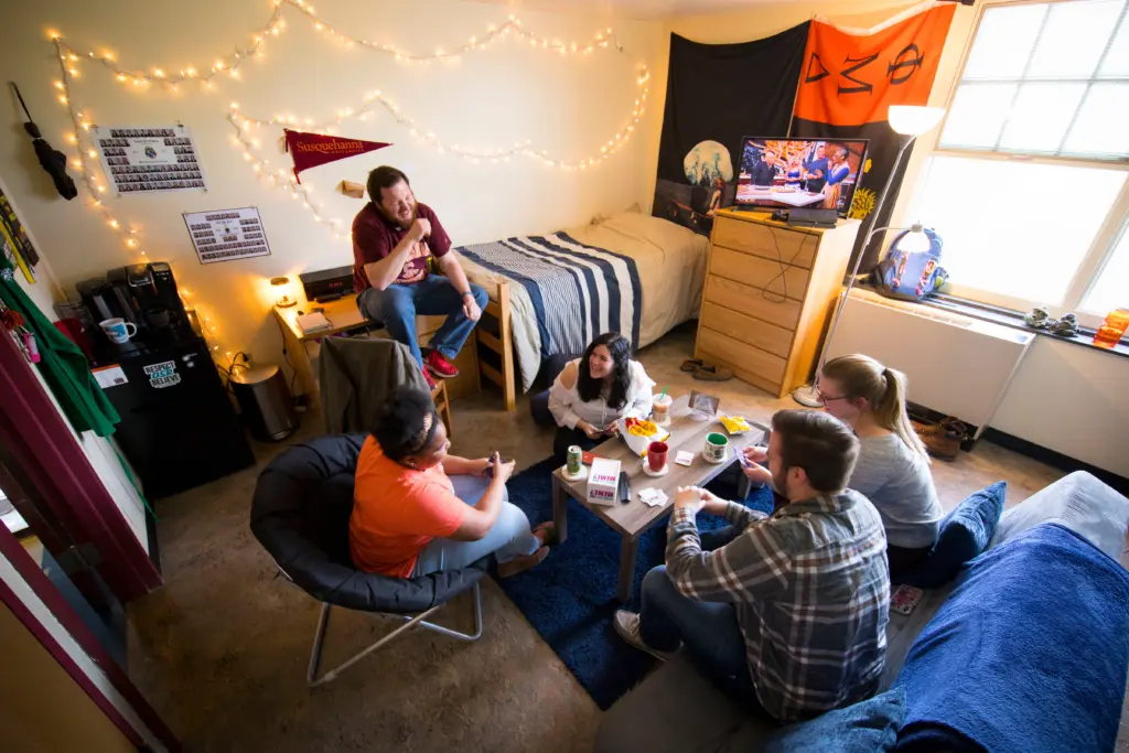 A group of five people are in a cozy upper-level housing dorm room. They sit around a small table, playing a game and eating snacks. The room is adorned with string lights, a flag on the wall, and a TV displaying a show.