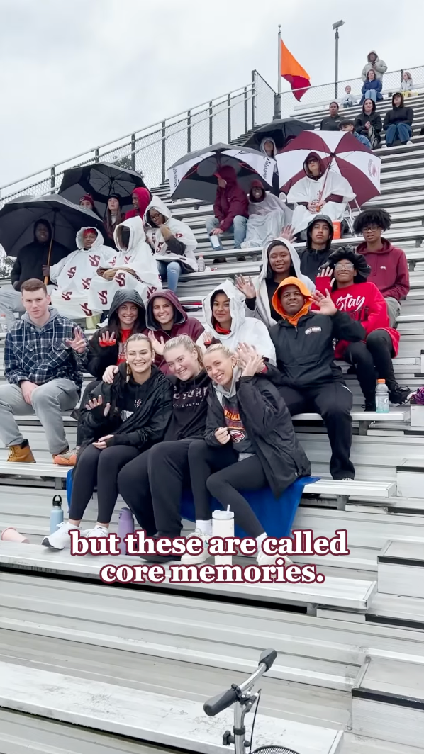 A group of people sit on bleachers at an outdoor event. Some hold umbrellas and wear jackets, indicating cold or rainy weather. Smiling and waving at the camera, they share core memories reminiscent of living on campus.
