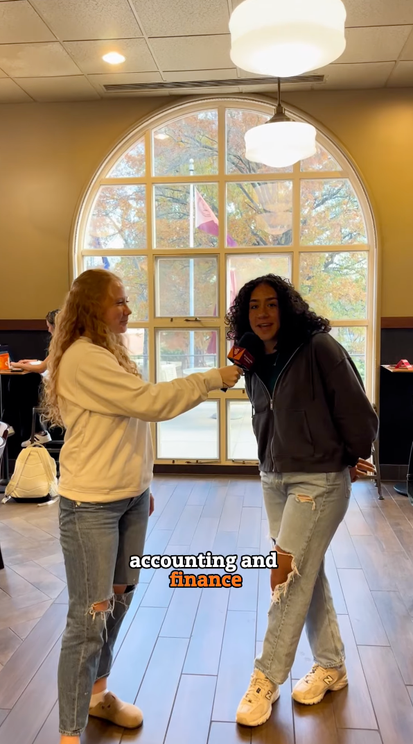 A person with curly hair is speaking into a microphone held by another person with long hair. They are indoors, standing in front of a large arched window with trees visible outside. The text reads, "academics and finance.