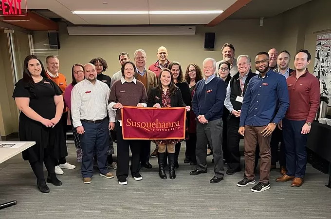 A group of about twenty alumni stands indoors, smiling at the camera. The central group holds a red Susquehanna University banner. The setting appears to be a meeting or event space with tables and posters.