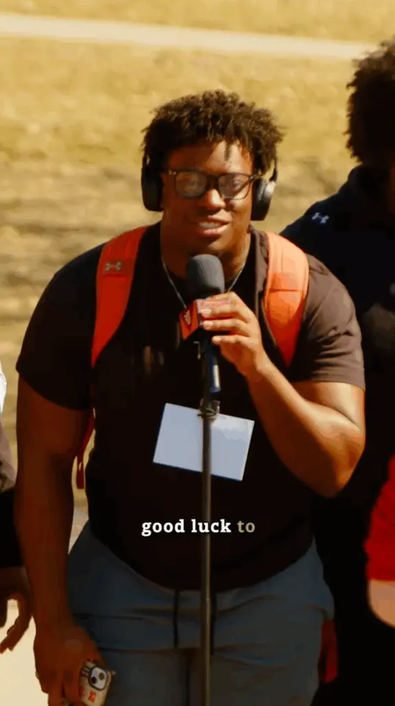 A person with headphones, glasses, and an orange backpack speaks into a microphone outdoors, capturing the essence of campus life. Wearing a black shirt, gray pants, and a name tag, they hold a phone as "good luck to" appears on the image.